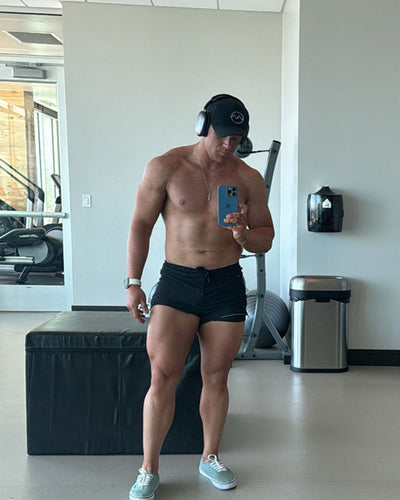 A shirtless man taking a mirror selfie in a gym, wearing black athletic shorts, a black baseball cap, over-ear headphones, and light blue sneakers.