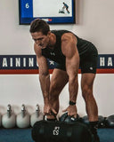 A muscular man in a black Under Armour athletic tank top and shorts bends over to lift a black 53 lb sandbag in a gym setting.