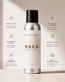 A bottle of MAKAI SUNLESS Airbrush Bronzing Mist in 'Kiss of Coconut Scent' is centered on a light beige background with stylized text callouts on both sides detailing its benefits. The bottle is metallic silver with a black cap and a cream-colored label featuring a subtle palm tree design.