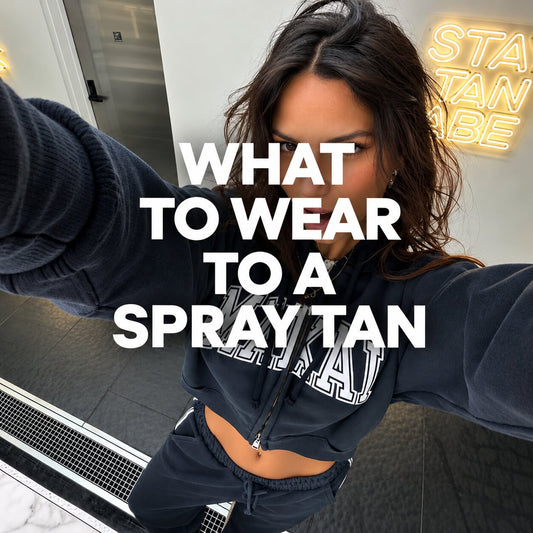 Woman taking a selfie in a spray tanning studio, wearing a dark cropped sweatshirt and matching joggers, with a spray tan booth and illuminated “Spray Tan” sign in the background.